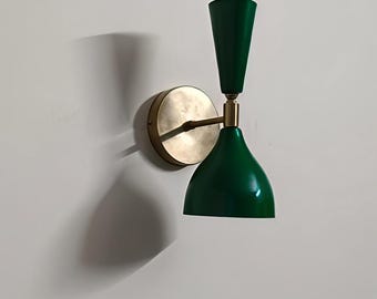 May include: A green and brass wall sconce with a conical shade and a cylindrical top. The sconce has a circular brass base and a curved arm. The fixture is mounted on a white wall.