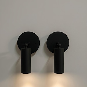May include: Two black wall sconces with a circular backplate and cylindrical light fixture. The sconces are mounted on a light-colored wall, with the light shining downwards. The sconces are a modern design.