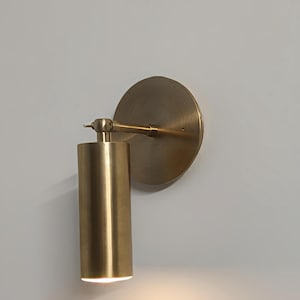 May include: A cylindrical brass wall sconce with a circular backplate. The sconce has a warm, golden hue and emits a soft light from its bottom. The design is minimalist and modern, suitable for interior lighting.