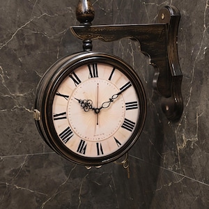 Op de afbeelding: Een dubbelzijdige wandklok met een donkerbruine, verweerde afwerking en een decoratieve muurbeugel. De wijzerplaat is gebroken wit met zwarte Romeinse cijfers en sierlijke zwarte metalen wijzers. Een kleine knop siert de bovenkant van de klok.