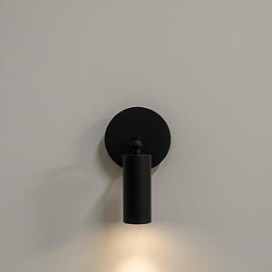 Puede incluir: Aplique de pared negro y moderno con una placa trasera circular y una lámpara cilíndrica. El aplique está montado en una pared blanca, y la luz brilla hacia abajo, creando un brillo cálido. Este diseño minimalista es adecuado para varios estilos de interior.