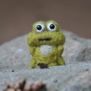 May include: A handmade, green felt frog figurine with large, black-pupiled eyes and a white chest. The frog is standing on a gray stone surface, with a blurred pinecone in the foreground. The frog is a whimsical, decorative item.