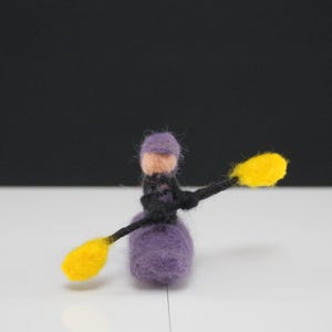 May include: A felted figure of a kayaker, featuring a purple body, a lavender hat, and a black outfit. The kayak paddle has yellow blades. The figure is set against a white surface and a black backdrop.