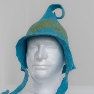 May include: A bright blue felted hat with a pointed, curled top and a wide brim. The hat features a soft, textured green patch across the front and includes two long, thin blue felted ties hanging down from the sides of the brim.