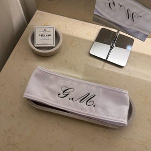 Op de afbeelding: Een witte badstof hoofdband geborduurd met de initialen "G.M." in zwart schrift. De hoofdband ligt op een bijpassend ovaal dienblad. Een klein doosje badzeep en een fles bodylotion zijn ook zichtbaar.