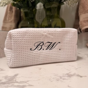 May include: White waffle-textured cosmetic bag with the embroidered initials "B.W." in black script. The bag is rectangular with a small bow on the side. The background includes green vases with flowers and a marble countertop.