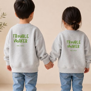Könnte beinhalten: Zwei hellgraue Sweatshirts mit Rundhalsausschnitt und dem grünen Schriftzug TROUBLE MAKER auf der Rückseite. Ein Sweatshirt trägt den Text Est. 2021, das andere Est. 2023. Beide Kleidungsstücke haben lange Ärmel und Rippbündchen, kombiniert mit blauen Jeans.