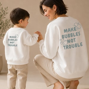 Könnte beinhalten: Zwei weiße langärmelige Sweatshirts mit dem Schriftzug "MAKE BUBBLES NOT TROUBLE" in blauen Buchstaben auf der Rückseite. Das Design enthält kleine kreisförmige Blasengrafiken um den Text herum. Die Sweatshirts haben einen Rundhalsausschnitt und gerippte Bündchen.