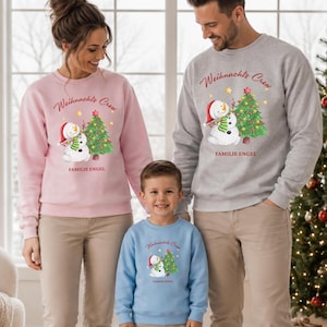 Könnte beinhalten: Ein hellrosa, ein hellgrauer und ein hellblauer Pullover, jeweils mit einer Grafik eines Schneemanns mit roter Mütze und grünem Schal neben einem geschmückten Weihnachtsbaum sowie dem Text "Weihnachts Crew" und "FAMILIE ENGEL" bedruckt.