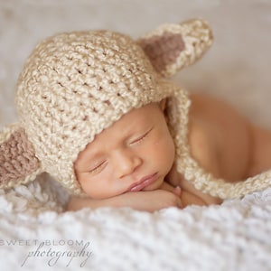 May include: A newborn baby wearing a crocheted cream-colored hat with brown ears, sleeping on a white fluffy blanket.