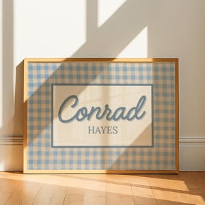 May include: A rectangular framed print featuring a light blue and white gingham pattern border surrounding a central cream-colored rectangle. The text Conrad is written in a blue script font above the word HAYES in a blue serif font.