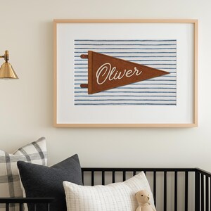 May include: A framed wall art print featuring a brown felt pennant with the name Oliver in white script, set against a blue and white striped background. The print is displayed in a light wood frame above a black crib with various pillows.