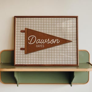 May include: A framed brown pennant with the name "Dawson" and the word "Hayes" in a cursive font. The pennant is brown and set against a beige and white checkered background. The frame is brown and the pennant is displayed on a green and beige shelf.