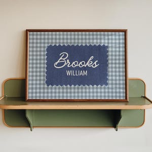 May include: A rectangular wooden frame containing a light blue and white gingham patterned background. Centered is a dark blue fabric patch with scalloped edges featuring the name Brooks in white script and WILLIAM in white block letters.
