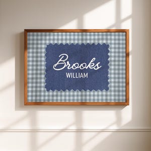 May include: A rectangular wooden frame containing a light blue and white gingham patterned background. Centered is a dark blue fabric patch with pinked edges featuring the white text "Brooks" in script and "WILLIAM" in a sans-serif font.