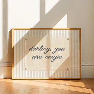 May include: A framed print featuring the phrase "darling, you are magic" in a script font. The print has a beige background with a striped border in light grey and white. The frame is a light wood colour.