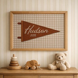Può includere: Un gagliardetto incorniciato con i nomi "Hudson" e "Jude" in materiale marrone simile alla pelle. Lo sfondo è un motivo a quadretti beige e bianco. Un elefante giocattolo di legno, un orsacchiotto e un giocattolo impilabile in legno sono su una cassettiera di legno.