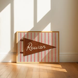 May include: A framed rectangular print featuring a brown triangular pennant with the name Rowan written in white cursive script. The pennant is centered over a background of vertical red and beige stripes, all enclosed in a light wood frame.