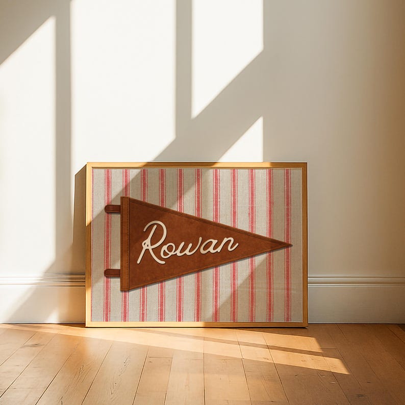 May include: A framed rectangular print featuring a brown triangular pennant with the name Rowan written in white cursive script. The pennant is centered over a background of vertical red and beige stripes, all enclosed in a light wood frame.