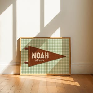 May include: A framed print featuring a brown pennant with the name "NOAH" and the name "thomas" in a serif font. The pennant is set against a green and white gingham pattern. The frame is a light brown color.