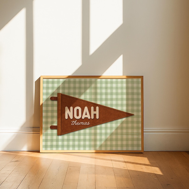 May include: A framed print featuring a brown pennant with the name "NOAH" and the name "thomas" in a serif font. The pennant is set against a green and white gingham pattern. The frame is a light brown color.