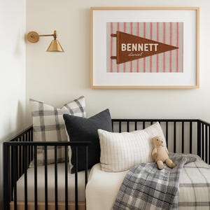 May include: A framed wall art print featuring a brown felt pennant with the name BENNETT and the name daniel printed in white. The pennant is set against a background of vertical red and white stripes, all enclosed in a light wood frame.