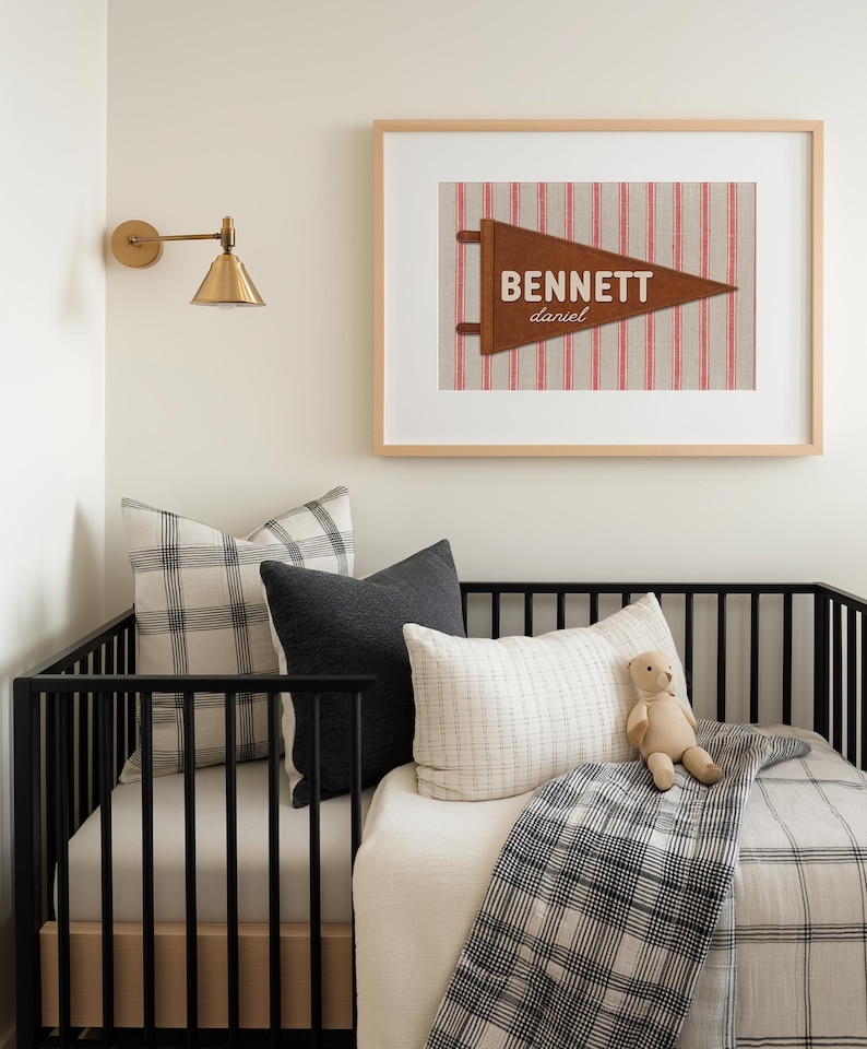 May include: A framed wall art print featuring a brown felt pennant with the name BENNETT and the name daniel printed in white. The pennant is set against a background of vertical red and white stripes, all enclosed in a light wood frame.