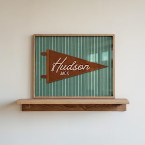 May include: A framed wall art piece featuring a brown felt pennant with the white text Hudson and Jack, centered on a green fabric background with thin white vertical pinstripes. The frame is made of light-colored wood and has a simple design.