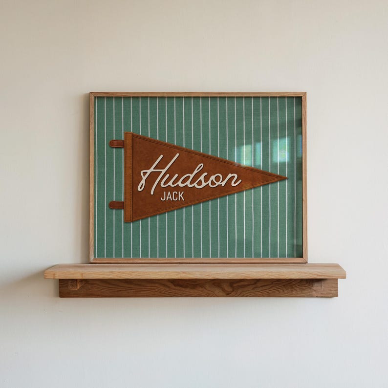 May include: A framed wall art piece featuring a brown felt pennant with the white text Hudson and Jack, centered on a green fabric background with thin white vertical pinstripes. The frame is made of light-colored wood and has a simple design.