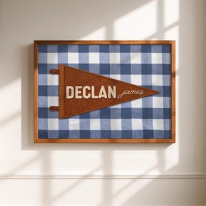 May include: A framed wall art piece featuring a blue and white gingham pattern. A brown pennant with the name "DECLAN james" is centered on the artwork. The frame is made of wood.