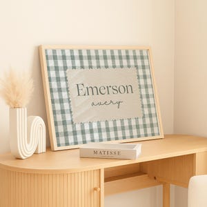 May include: A framed wall art print featuring a sage green and white gingham pattern background with a central beige rectangular patch. The text Emerson is printed in a dark gray serif font above the name avery in a dark gray script font.