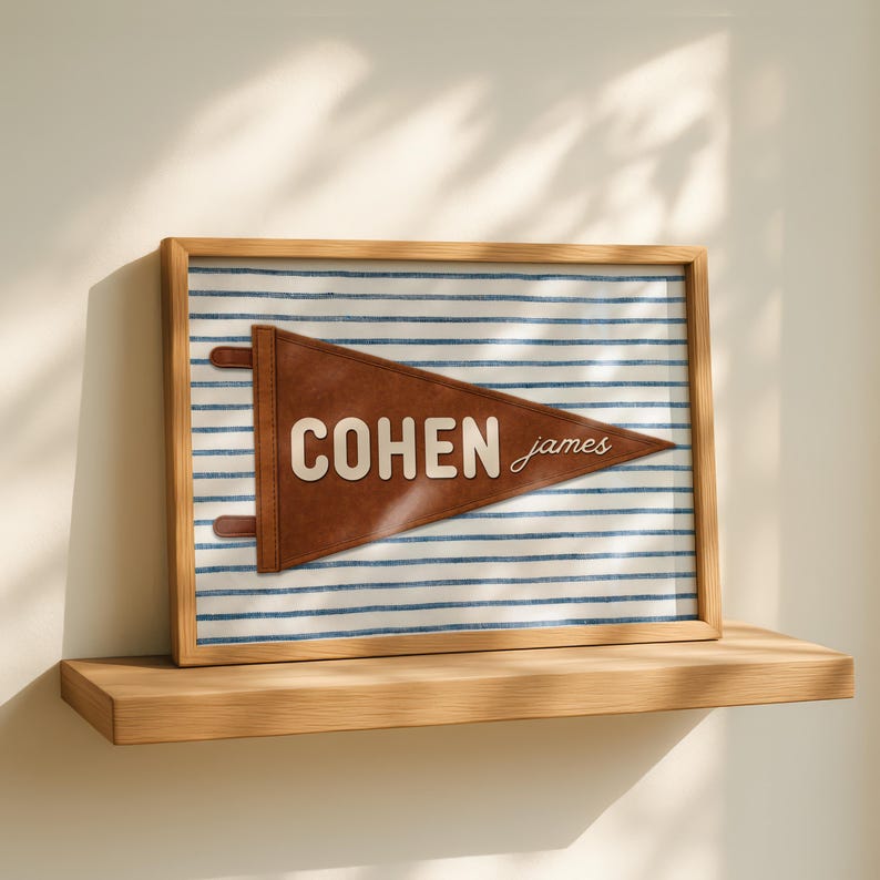 May include: A framed wall art piece featuring a brown leather pennant with the name COHEN JAMES in white lettering. The pennant is mounted on a white background with thin horizontal blue stripes, all enclosed in a light wood frame.