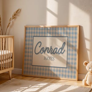 May include: A rectangular framed wall art piece featuring a light blue and beige gingham pattern background. A central cream-colored rectangle contains the name Conrad in a large blue script font, with the name HAYES in a smaller blue sans-serif font.