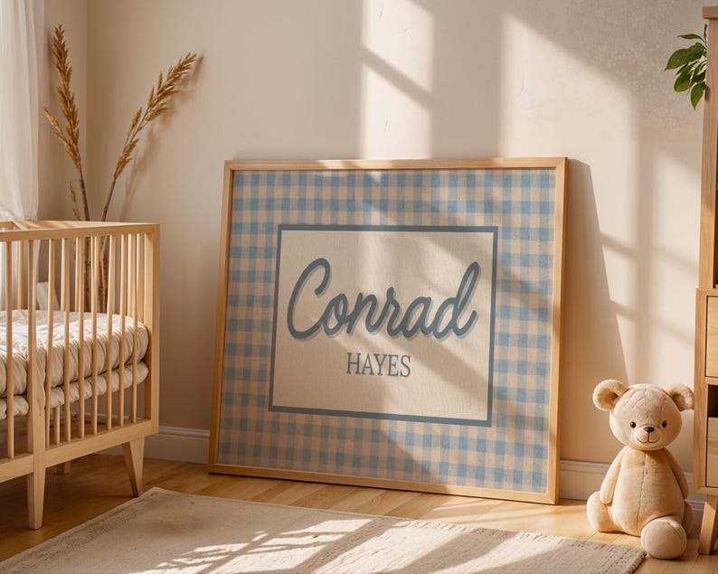 May include: A rectangular framed wall art piece featuring a light blue and beige gingham pattern background. A central cream-colored rectangle contains the name Conrad in a large blue script font, with the name HAYES in a smaller blue sans-serif font.