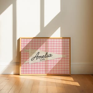 May include: A framed rectangular print featuring a white pennant flag centered on a pink and white gingham background. The pennant displays the name Amelia in a black cursive script with the word JUNE printed in black block letters underneath.