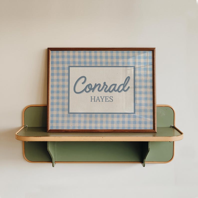 May include: A rectangular framed print featuring a light blue and white gingham pattern border surrounding a central white panel. The text "Conrad" is written in a blue cursive script above the word "HAYES" in a blue sans-serif font.