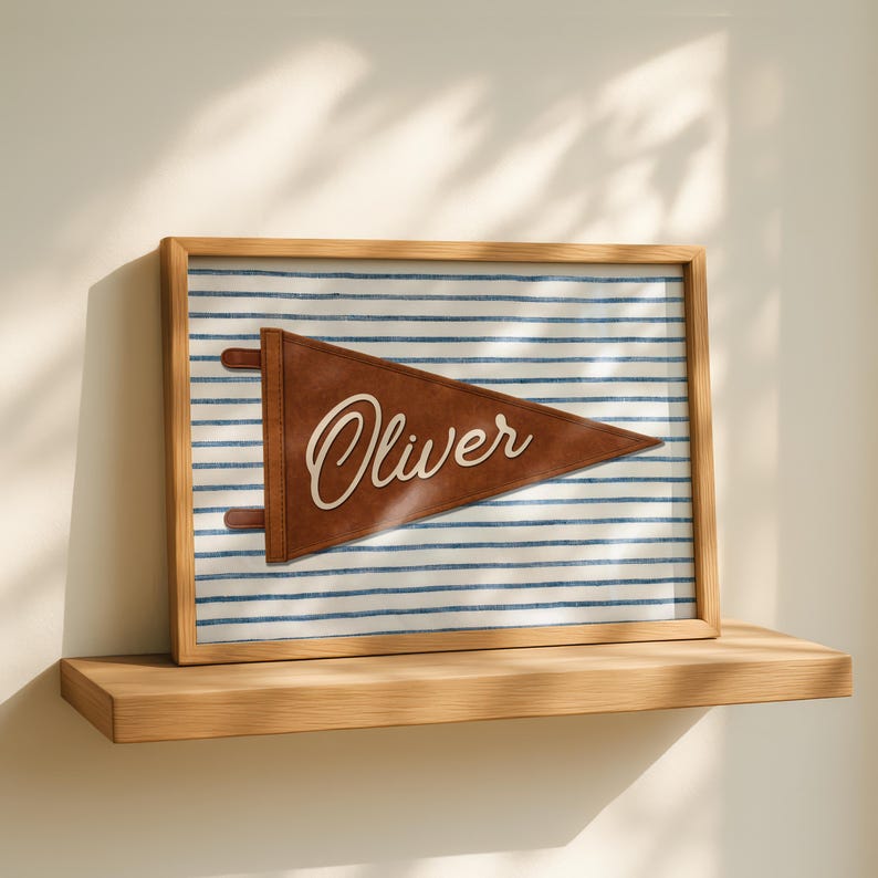 May include: A framed pennant with the name "Oliver" in white script. The pennant is brown and set against a blue and white striped background. The frame is made of light-colored wood, and the artwork is displayed on a wooden shelf.