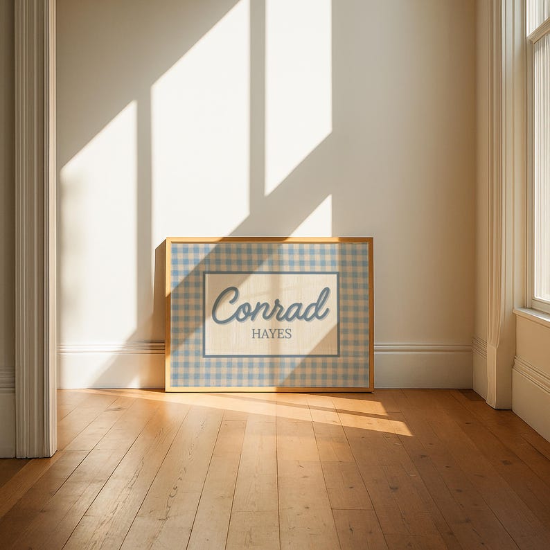 May include: A rectangular framed print featuring a light blue and white gingham pattern border surrounding a central cream-colored rectangle. The text Conrad is written in a blue script font above the word HAYES in a blue serif font.