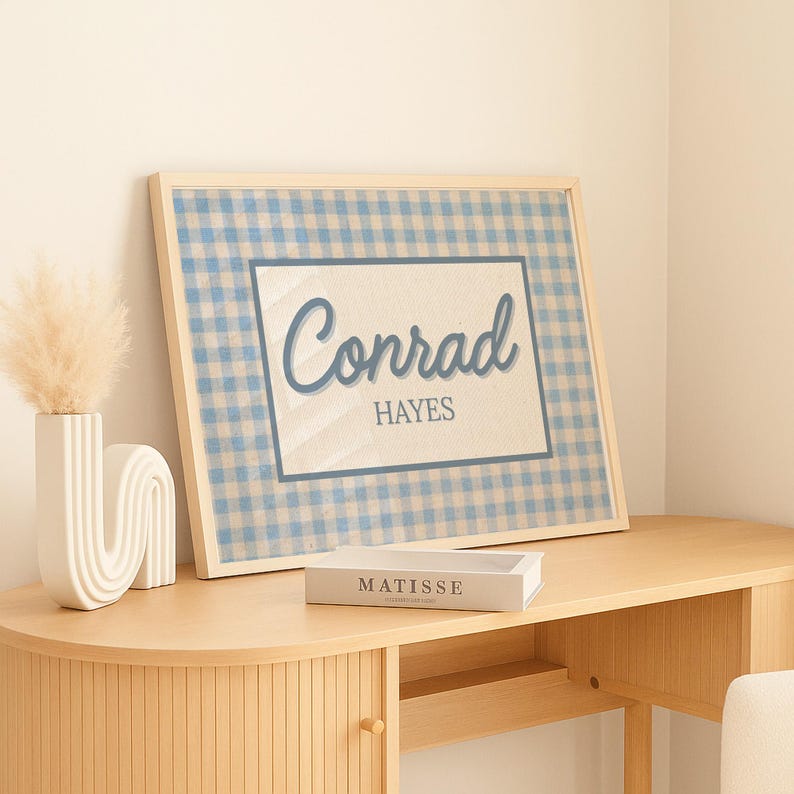 May include: A framed rectangular print featuring a light blue and white gingham pattern background. The center contains a white rectangular panel with a dark blue border, displaying the name Conrad in script and HAYES in block letters.