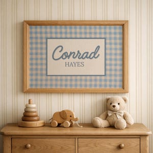 May include: A rectangular wooden frame containing a print with a light blue and white gingham pattern background. A central white rectangle features the name Conrad in a blue script font above the name HAYES in a blue block font.