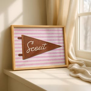 May include: A framed wall art piece featuring a brown felt pennant with the word Scout in white cursive script. The pennant is centered on a background of horizontal light pink and white stripes with a textured fabric appearance.