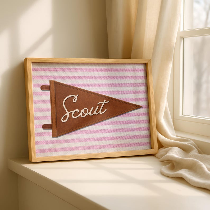 May include: A framed wall art piece featuring a brown felt pennant with the word Scout in white cursive script. The pennant is centered on a background of horizontal light pink and white stripes with a textured fabric appearance.