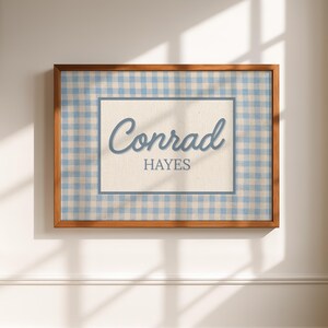 May include: A rectangular wall art piece in a wooden frame featuring a light blue and white gingham pattern border. The center contains a cream-colored rectangular panel with the name Conrad Hayes printed in blue, with Conrad in script.