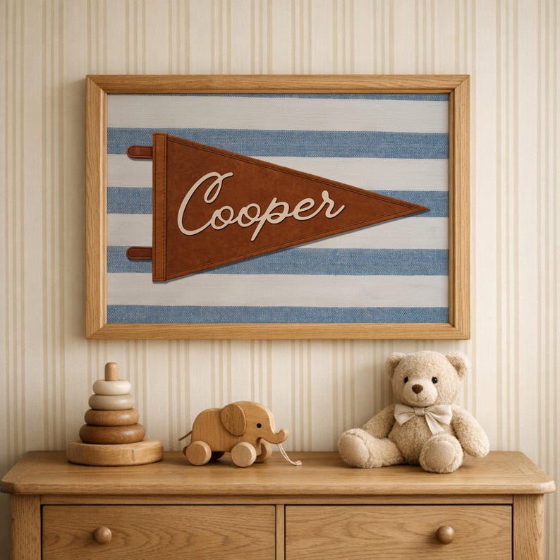 May include: A framed wall art piece with a brown pennant that reads "Cooper" over a blue and white striped background. A wooden toy elephant, a stacking ring toy, and a teddy bear are on a wooden dresser.