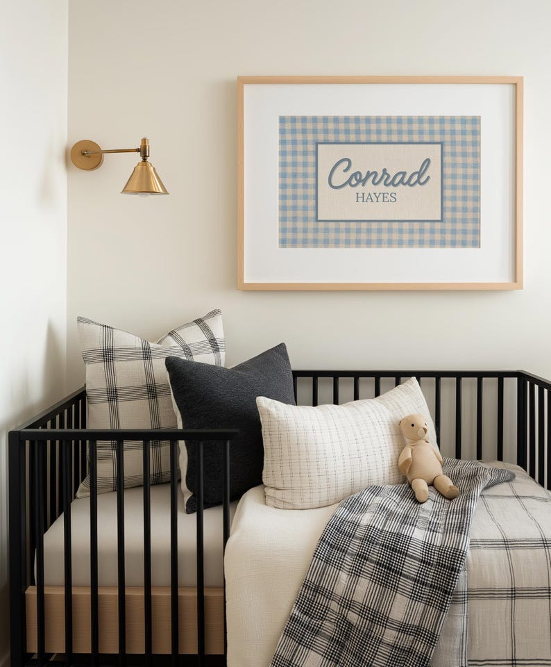 May include: A framed wall art print featuring a blue and white gingham pattern background with a central rectangular border containing the name Conrad Hayes in blue script and block lettering, displayed above a black crib with pillows.