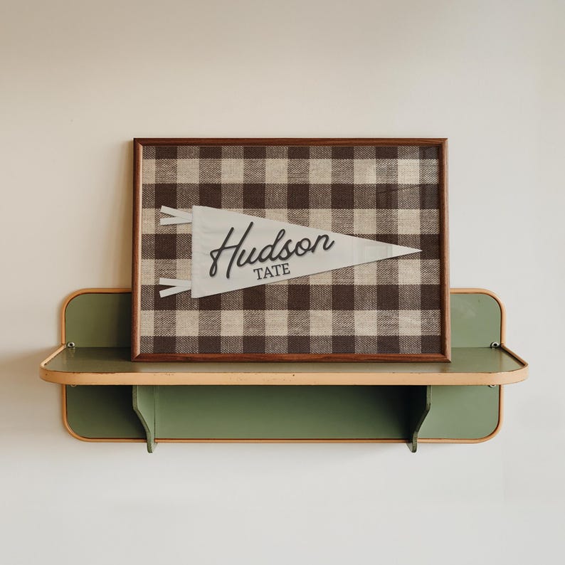 May include: A rectangular framed wall art piece featuring a brown and white buffalo check fabric background. Centered on the fabric is a white felt pennant with two small ties on the left side and the text Hudson TATE in black lettering.