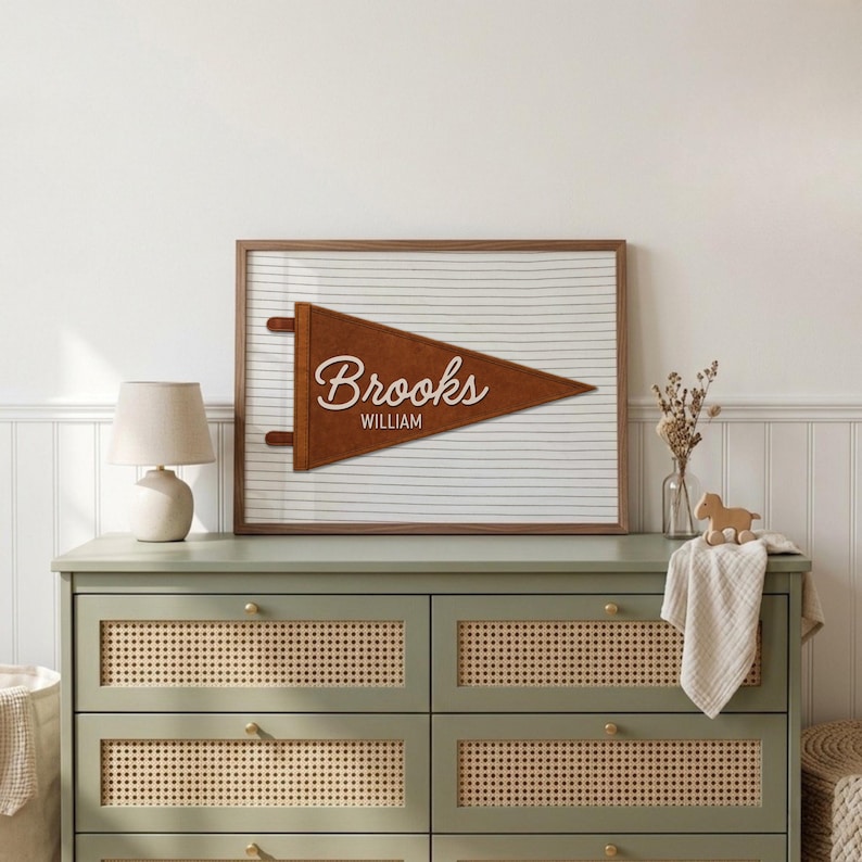 May include: A framed wall art piece featuring a brown felt pennant with the name Brooks in white script and the name William in white block letters, set against a white background with thin horizontal black lines.