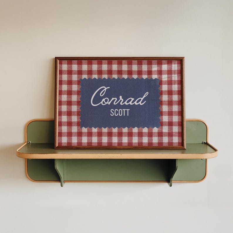 May include: A rectangular wooden frame containing a red and white gingham patterned background with a centered blue scalloped-edge rectangle featuring the white text Conrad in script and the white text SCOTT in a sans-serif font.