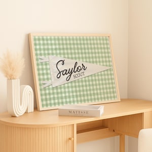May include: A rectangular framed wall art piece featuring a light green and white gingham check pattern background. Centered on the print is a white triangular pennant with the text Saylor SCOUT written in a dark, cursive script font.