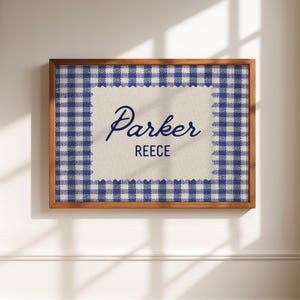 May include: A rectangular wall art piece in a wooden frame featuring a blue and white gingham border surrounding a central cream-colored fabric panel. The text 'Parker' is written in a blue script font above the word 'REECE' in blue block letters.